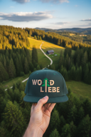 Aus der Ich-Perspektive hält eine Hand eine dunkelgrüne Kappe mit dem "WOID LIEBE"-Schriftzug in den Vordergrund. Im Hintergrund erstreckt sich eine weite, malerische Landschaft aus dichten Wäldern und sanften Hügeln, die in das goldene Licht der Abendsonne getaucht sind.