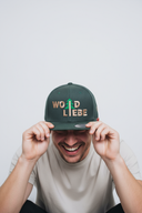 Ein Mann lacht ausgelassen, während er vor einem hellgrauen Studiohintergrund sitzt und sich mit beiden Händen eine dunkelgrüne Kappe mit dem goldfarbenen Schriftzug "WOID LIEBE" ins Gesicht zieht.