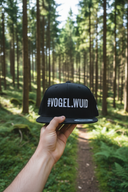 Aus der Ich-Perspektive hält eine Hand eine schwarze Kappe mit dem "#VOGEL.WUID"-Schriftzug in den Vordergrund. Im Hintergrund erstreckt sich ein sonnendurchfluteter Waldweg, der von hohen Nadelbäumen und grünem Farn gesäumt ist.