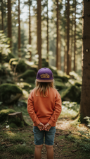 Ein Mädchen mit langen blonden Haaren, von hinten fotografiert, steht mit hinter dem Rücken verschränkten Händen in einem moosbewachsenen, sonnendurchfluteten Wald. Sie trägt einen orangen Pullover und eine lila Kappe mit dem gelben Schriftzug "Wuide HENA".