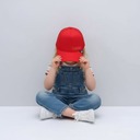 Ein Kind in einer Jeans-Latzhose sitzt im Schneidersitz vor einem hellgrauen Studiohintergrund und hält sich mit beiden Händen eine rote Trucker-Kappe mit der Ton-in-Ton-Aufschrift "LAUSDEANDL" vor das Gesicht.