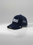 Ein professionelles Produktfoto einer dunkelblauen Trucker-Kappe. Auf der Vorderseite befindet sich der erhabene weiße Schriftzug "Bazi". Die Kappe steht auf einer spiegelnden Oberfläche vor einem hellen Studiohintergrund, was eine deutliche Reflexion erzeugt.