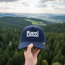Eine Hand hält eine dunkelblaue Kappe mit dem weißen Schriftzug "Bazi" in den Vordergrund. Im Hintergrund erstreckt sich eine weite, malerische Landschaft aus dichten Wäldern und sanften Hügeln.