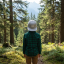 Ein Junge, von hinten fotografiert, steht auf einem Waldweg und blickt durch eine Lücke zwischen den hohen Nadelbäumen auf einen Berggipfel. Er trägt ein grün-schwarzes Karohemd und eine graue Kappe mit der Aufschrift "Bazi".