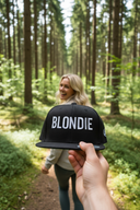 Aus der Ich-Perspektive hält eine Hand eine schwarze Kappe mit dem weißen Schriftzug "BLONDIE" in die Kamera. Im unscharfen Hintergrund geht eine lachende Frau mit langen blonden Haaren auf einem sonnendurchfluteten Waldweg.