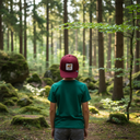 Ein kleiner Junge, von hinten fotografiert, steht in einem moosbewachsenen, sonnendurchfluteten Nadelwald. Er trägt ein grünes T-Shirt und eine weinrote Kappe mit einem Fuchs-Aufnäher.
