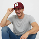 Ein Mann mit einem breiten Lächeln sitzt vor einem hellgrauen Studiohintergrund und berührt den Schirm seiner weinroten Kappe. Auf der Kappe befindet sich ein Aufnäher mit der Abbildung eines Fuchses und dem Schriftzug "FUX".