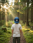 Ein kleiner Junge, von hinten fotografiert, steht auf einem Waldweg, während Sonnenstrahlen durch die hohen Bäume fallen und eine zauberhafte Atmosphäre schaffen. Er trägt ein helles T-Shirt und eine blaue Kappe mit einem Hahn-Aufnäher.