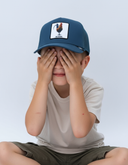 Ein Junge sitzt im Schneidersitz vor einem hellgrauen Studiohintergrund und hält sich die Hände vor die Augen. Er trägt eine blaue Trucker-Kappe mit einem quadratischen Aufnäher, der einen Hahn und den Schriftzug "GICKERL" zeigt.