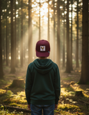 Ein kleiner Junge, von hinten fotografiert, steht auf einem Waldweg, während Sonnenstrahlen durch die hohen Bäume fallen und eine zauberhafte Atmosphäre schaffen. Er trägt einen grünen Kapuzenpullover und eine weinrote Kappe mit einem Huhn-Aufnäher.