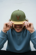Ein Mann mit einem breiten Lächeln sitzt vor einem hellgrauen Hintergrund und zieht sich mit beiden Händen eine olivgrüne Kappe mit gelbem Schirm ins Gesicht. Auf der Kappe ist der Kopf eines Hirsches gestickt.