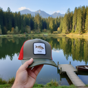 Aus der Ich-Perspektive hält eine Hand eine Kappe mit einem Karpfen-Aufnäher in den Vordergrund. Im Hintergrund liegt ein ruhiger Bergsee, in dem sich die umliegenden Nadelwälder und Berggipfel spiegeln. Ein kleiner Holzsteg führt auf das Wasser.