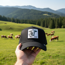 Aus der Ich-Perspektive hält eine Hand eine schwarze Kappe mit einem Kuhkopf-Aufnäher in den Vordergrund. Im Hintergrund grast eine Herde Kühe auf einer saftig grünen Weide vor einer Kulisse aus dichten Wäldern und Hügeln.