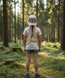 Ein Mädchen mit einem geflochtenen Zopf, von hinten fotografiert, steht in einem moosbewachsenen, sonnendurchfluteten Wald. Sie trägt ein helles T-Shirt und eine beige Kappe mit einem Eichhörnchen-Aufnäher.