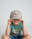 Ein kleiner Junge sitzt im Schneidersitz vor einem hellgrauen Studiohintergrund und zieht sich mit beiden Händen eine beige Kappe über die Augen. Die Kappe hat einen quadratischen Aufnäher, der ein Eichhörnchen und den Schriftzug "OACHKATZL" zeigt.