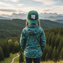 Ein Mädchen mit Zöpfen, von hinten fotografiert, steht auf einem Aussichtspunkt und blickt auf eine beeindruckende Berglandschaft. Sie trägt eine gemusterte grüne Jacke und eine grüne Kappe mit einem Schaf-Motiv.