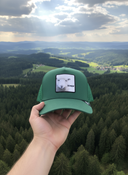 Aus der Ich-Perspektive hält eine Hand eine grüne Kappe mit einem Schaf-Aufnäher in den Vordergrund. Im Hintergrund erstreckt sich eine weite, malerische Landschaft aus dichten Wäldern und sanften Hügeln unter einem leicht bewölkten Himmel.