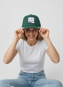 Eine Frau lacht ausgelassen, während sie vor einem hellgrauen Studiohintergrund eine grüne Kappe mit beiden Händen über ihre Augen zieht. Auf der Kappe ist ein Aufnäher mit einem Schaf und dem Schriftzug "SCHOOF" zu sehen.