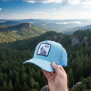 Aus der Ich-Perspektive hält eine Hand eine hellblaue Kappe mit einem Wolf-Aufnäher in den Vordergrund. Im Hintergrund erstreckt sich eine weite, malerische Landschaft aus dichten Wäldern, nebligen Tälern und felsigen Hügeln.