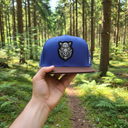 Aus der Ich-Perspektive hält eine Hand eine blau-braune Kappe mit einem gestickten Wildschweinkopf in den Vordergrund. Im Hintergrund erstreckt sich ein sonnendurchfluteter Waldweg, der von hohen Nadelbäumen und grünem Farn gesäumt ist.