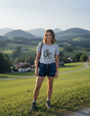 Eine lächelnde Frau mit schulterlangem, braunem Haar steht auf einem saftig grünen Hügel in einer alpinen Landschaft. Sie trägt ein graues T-Shirt mit Aufdruck, blaue Jeans-Shorts und Wanderstiefel und hat einen Rucksack auf. Im Hintergrund erstreckt sich ein malerisches Tal mit verstreuten Häusern und sanften Hügeln, die zu einer Bergkette in der Ferne führen.