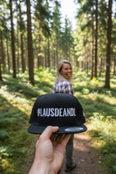 Aus der Ich-Perspektive hält eine Hand eine schwarze Kappe mit dem weißen Schriftzug "#LAUSDEANDL" in die Kamera. Im unscharfen Hintergrund geht eine Frau auf einem Waldweg und blickt über die Schulter zurück, wobei sie spielerisch die Zunge herausstreckt.