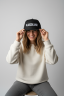 Eine lächelnde Frau sitzt auf einem Hocker vor einem hellgrauen Studiohintergrund. Sie setzt sich gerade eine schwarze Kappe mit dem weißen Schriftzug "#LAUSDEANDL" auf und blickt dabei freundlich in die Kamera.