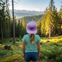 Ein Mädchen mit einem blonden Pferdeschwanz, von hinten fotografiert, steht in einem sonnigen Nadelwald. Es trägt eine lila Kappe mit rotem Blumenschriftzug und blickt auf eine ferne Bergkette.