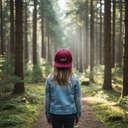 Ein Mädchen mit langen blonden Haaren, von hinten fotografiert, steht auf einem Waldweg, während Sonnenstrahlen durch die hohen Bäume fallen und eine zauberhafte Atmosphäre schaffen. Sie trägt eine Jeansjacke und eine weinrote Kappe mit der weißen Aufschrift "Lausdeandl".