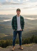 Ein lächelnder junger Mann steht mit den Händen in den Hosentaschen auf einem Aussichtspunkt in den Bergen. Er trägt ein grün kariertes Hemd und eine grüne Kappe mit Lederaufnäher. Im Hintergrund erstreckt sich eine weite Landschaft aus bewaldeten Hügeln und nebelverhangenen Tälern.