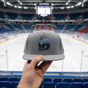 Aus der Ich-Perspektive hält eine Hand eine graue Kappe mit dem "Marcel Brandt"-Logo in den Vordergrund. Im Hintergrund erstreckt sich der Blick über eine leere Eishockey-Arena mit frisch präpariertem Eis, blauen Sitzen und einer großen Anzeigetafel.