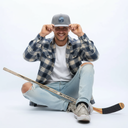 Ein Eishockeyspieler, vermutlich Marcel Brandt, sitzt mit einem breiten Lächeln im Schneidersitz vor einem weißen Studiohintergrund. Vor ihm liegt ein Eishockeyschläger, während er sich mit beiden Händen eine graue Kappe mit seinem persönlichen Logo aufsetzt.