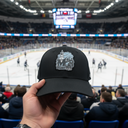Aus der Perspektive eines Zuschauers hält eine Hand eine schwarze Sportkappe mit dem "Marcel Brandt"-Logo in den Vordergrund. Im unscharfen Hintergrund findet auf der Eisfläche einer gut besuchten Arena ein Eishockeyspiel statt.