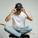 Ein Eishockeyspieler, vermutlich Marcel Brandt, sitzt mit einem breiten Lächeln im Schneidersitz auf einem Hocker. Er balanciert einen Eishockeyschläger auf seinen Knien, während er sich mit beiden Händen eine schwarze Sportkappe mit seinem persönlichen Logo aufsetzt.