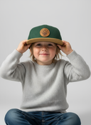 1. For the file vogel-wuid-Muaehlbauer-Brauerei-Cap-3-Kids-studio.jpg: Ein Junge mit Sommersprossen sitzt im Schneidersitz vor einem hellgrauen Studiohintergrund. Er setzt sich mit beiden Händen eine grüne Kappe mit braunem Schirm und einem runden Lederaufnäher auf und blickt dabei freundlich in die Kamera.