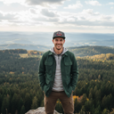 Ein lächelnder Mann steht mit den Händen in den Hosentaschen auf einem Felsen vor einer weiten Landschaft aus herbstlich gefärbten Wäldern und Hügeln. Er trägt eine grüne Jacke und eine graue Kappe mit einem "Mühlbauer"-Brauerei-Logo.