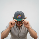Ein Mann mit einem breiten Lächeln sitzt vor einem hellgrauen Studiohintergrund und zieht sich mit beiden Händen eine grau-grüne Kappe mit einem bunten Brauerei-Logo ins Gesicht.