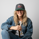 Eine junge Frau in einer Jeansjacke sitzt vor einem hellgrauen Studiohintergrund und lächelt freundlich in die Kamera. Sie trägt eine grau-grüne Kappe mit einem "Mühlbauer"-Brauerei-Logo und hält eine Flasche Bier in der Hand.