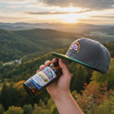 Aus der Ich-Perspektive hält eine Hand eine Bierflasche, auf der eine grau-grüne Kappe mit Brauerei-Logo balanciert. Im Hintergrund erstreckt sich eine weite, hügelige Waldlandschaft im Licht der untergehenden Sonne.