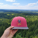 Aus der Ich-Perspektive hält eine Hand eine pinke Kappe mit dem "Oimi"-Logo in den Vordergrund. Im Hintergrund erstreckt sich eine weite, malerische Landschaft aus dichten Wäldern und sanften Hügeln unter einem leicht bewölkten Himmel.