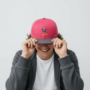 Ein Mann mit einem breiten Lächeln sitzt vor einem hellgrauen Studiohintergrund und zieht sich mit beiden Händen eine pinke Kappe mit grauem Schirm und "Oimi"-Aufdruck ins Gesicht.