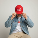 Ein Mann sitzt vor einem hellgrauen Studiohintergrund und zieht sich mit beiden Händen eine rote Kappe mit der weißen Aufschrift "REGIONAL STAR" und einem "Rewe"-Logo ins Gesicht.