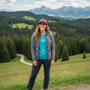 Eine lächelnde Frau in Wanderkleidung steht auf einer blühenden Bergwiese. Sie trägt eine graue Fleecejacke, ein leuchtend blaues T-Shirt und eine grau-rote Kappe. Im Hintergrund erstreckt sich eine weite Landschaft aus dichten Wäldern, sanften Hügeln und einer beeindruckenden Bergkette am Horizont. Ein Feldweg schlängelt sich durch die grünen Hänge.