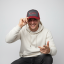 Ein Mann mit einem breiten, freundlichen Lächeln sitzt vor einem hellgrauen Studiohintergrund. Er trägt einen hellen Kapuzenpullover und berührt mit einer Hand den Schirm seiner grau-roten Kappe mit der Aufschrift "Xundheit", während er mit der anderen Hand spielerisch in Richtung Kamera zeigt.