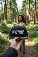 Aus der Ich-Perspektive hält eine Hand eine schwarze Kappe mit dem weißen Schriftzug "BROWNIE" in die Kamera. Im unscharfen Hintergrund geht eine lachende Frau mit langen braunen Haaren auf einem sonnendurchfluteten Waldweg.