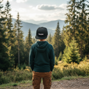 Ein Junge, von hinten fotografiert, steht mit den Händen in den Hosentaschen auf einem Aussichtspunkt und blickt auf eine weite Landschaft aus Nadelwäldern und Bergen. Er trägt einen grünen Kapuzenpullover und eine schwarze Kappe mit der weißen Aufschrift "Lausbua".