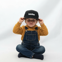 Ein kleiner Junge in Jeans-Latzhose und einem senfgelben Kapuzenpullover sitzt im Schneidersitz vor einem hellgrauen Studiohintergrund. Er setzt sich mit beiden Händen eine schwarze Kappe mit dem weißen Schriftzug "Lausbua" auf und blickt dabei direkt in die Kamera.