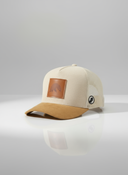 Ein professionelles Produktfoto einer beigen Trucker-Kappe mit braunem Wildlederschirm. Die Cord-Vorderseite ziert ein quadratischer Lederaufnäher mit einem geprägten Bergmotiv. Die Kappe steht auf einer spiegelnden Oberfläche vor einem hellen Studiohintergrund und erzeugt eine deutliche Reflexion.