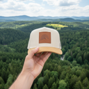 Aus der Ich-Perspektive hält eine Hand eine beige Kappe mit einem Lederaufnäher mit Bergmotiv in den Vordergrund. Im Hintergrund erstreckt sich eine weite, malerische Landschaft aus dichten Wäldern und sanften Hügeln.