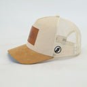 Eine beige Trucker-Kappe, fotografiert in der Seitenansicht vor einem weißen Hintergrund. Diese Ansicht zeigt deutlich die strukturierte Cord-Vorderseite, den braunen Wildlederschirm, den Lederaufnäher mit Bergmotiv und den hellen Mesh-Einsatz am Hinterkopf.